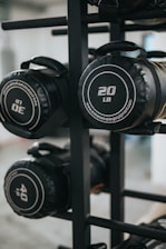 Several cylindrical fitness sandbags are stored on a black metal rack. The visible sandbags are labeled with different weights such as 20, 30, and 40 pounds and feature handles for easy carrying.