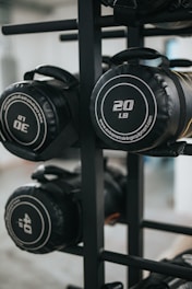 Several cylindrical fitness sandbags are stored on a black metal rack. The visible sandbags are labeled with different weights such as 20, 30, and 40 pounds and feature handles for easy carrying.