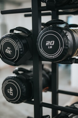 Several cylindrical fitness sandbags are stored on a black metal rack. The visible sandbags are labeled with different weights such as 20, 30, and 40 pounds and feature handles for easy carrying.
