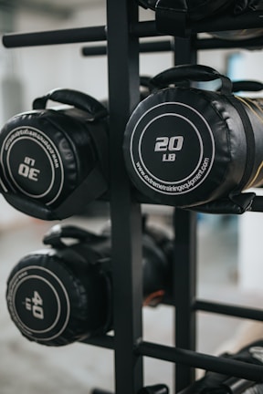 Several cylindrical fitness sandbags are stored on a black metal rack. The visible sandbags are labeled with different weights such as 20, 30, and 40 pounds and feature handles for easy carrying.