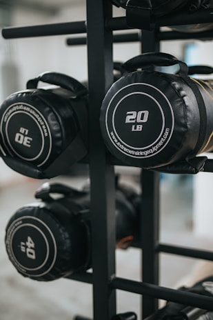 Several cylindrical fitness sandbags are stored on a black metal rack. The visible sandbags are labeled with different weights such as 20, 30, and 40 pounds and feature handles for easy carrying.