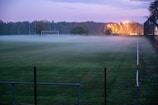 A football field is shrouded in mist during twilight, with dim lighting coming from distant street lamps. The field is surrounded by a wire fence, and the goal posts are visible through the fog. Trees line the horizon, adding a touch of nature to the serene atmosphere.