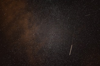 A night sky filled with numerous stars and a faint streak of light, possibly a meteor or satellite trail, crossing the frame diagonally. The sky has areas with small clouds or nebula-like formations.
