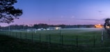 A football field with green grass is surrounded by a metal fence. The scene is set during twilight with a pastel sky blending blue and pink hues. There are trees in the background, and the field is empty with goalposts visible. Distant lights glow warmly against the evening sky, contributing to a serene atmosphere.