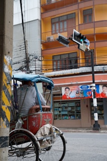 A person is sitting on a traditional rickshaw parked on a city street. The rickshaw has a red body with an old and worn appearance, covered by a blue canopy. The background shows a busy urban scene with a building that features a mix of orange and beige colors. Below the building, there is a shop with advertisements that include images of people. Above the street, a traffic light with green lights is visible, and there are numerous electrical wires overhead.