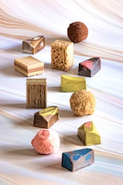An assortment of uniquely shaped and colored chocolates, each with distinct textures and designs, arranged on a swirling pastel background. The chocolates include geometric shapes, truffles, and textured pieces.