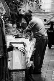 A person stands beside a stall or shop, surrounded by a variety of items including papers, packaging materials, and potted plants. The setting appears to be indoors or under a covered area with a focus on an industrial or commercial environment.