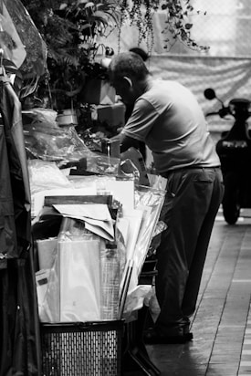 A person stands beside a stall or shop, surrounded by a variety of items including papers, packaging materials, and potted plants. The setting appears to be indoors or under a covered area with a focus on an industrial or commercial environment.