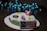 A cake decorated with fondant figures resembling characters and items from a video game, including a red and white mushroom, a yellow star, and a bob-omb. A fondant game controller is attached to the side of the cake. Blue LED string lights create a bokeh effect in the background.