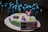A cake decorated with fondant figures resembling characters and items from a video game, including a red and white mushroom, a yellow star, and a bob-omb. A fondant game controller is attached to the side of the cake. Blue LED string lights create a bokeh effect in the background.