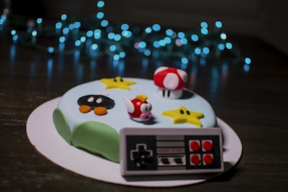 A cake decorated with fondant figures resembling characters and items from a video game, including a red and white mushroom, a yellow star, and a bob-omb. A fondant game controller is attached to the side of the cake. Blue LED string lights create a bokeh effect in the background.