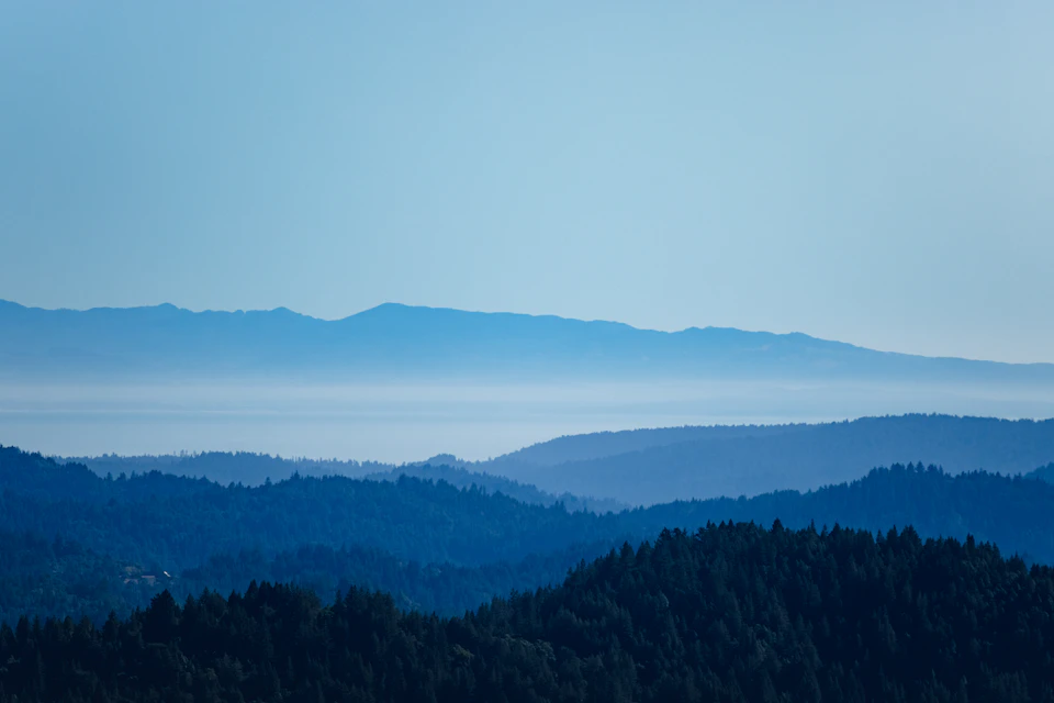 The image depicts a breathtaking view of a mountain range under a clear blue sky. The mountains, painted in various shades of blue and green, stretch across the horizon, creating a sense of depth and vastness. The perspective is from a high vantage point, looking down upon the mountains, which enhances their grandeur. In the foreground, there are trees that add a touch of life to the otherwise barren landscape. The image does not contain any discernible text or countable objects, making it an open invitation for viewers to imagine and interpret the scene in their own unique way.

The colors used in the image are predominantly blue and green, reflecting the natural beauty of the mountains and trees. The sky above is a clear blue, providing a striking contrast against the greenery below. Overall, the image presents a serene and awe-inspiring landscape that captures the essence of nature's grandeur.