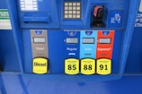 A gas station pump panel featuring four fuel options: Diesel, Regular, Extra, and Supreme+, each with distinct colors and labels. The panel includes pricing displays and a card payment slot. Each fuel option has a yellow button indicating the octane rating. The panel is predominantly blue with some black and red elements.