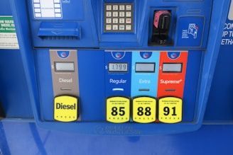 A gas station pump panel featuring four fuel options: Diesel, Regular, Extra, and Supreme+, each with distinct colors and labels. The panel includes pricing displays and a card payment slot. Each fuel option has a yellow button indicating the octane rating. The panel is predominantly blue with some black and red elements.