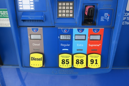 A gas station pump panel featuring four fuel options: Diesel, Regular, Extra, and Supreme+, each with distinct colors and labels. The panel includes pricing displays and a card payment slot. Each fuel option has a yellow button indicating the octane rating. The panel is predominantly blue with some black and red elements.