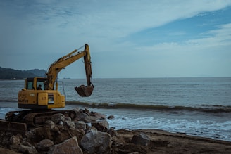 A yellow excavator is positioned on a rocky shoreline next to the ocean. The machine sits atop large stones, with its arm and bucket extended. The sea is calm, with small waves gently reaching the shore, and the sky is partly cloudy with patches of blue.