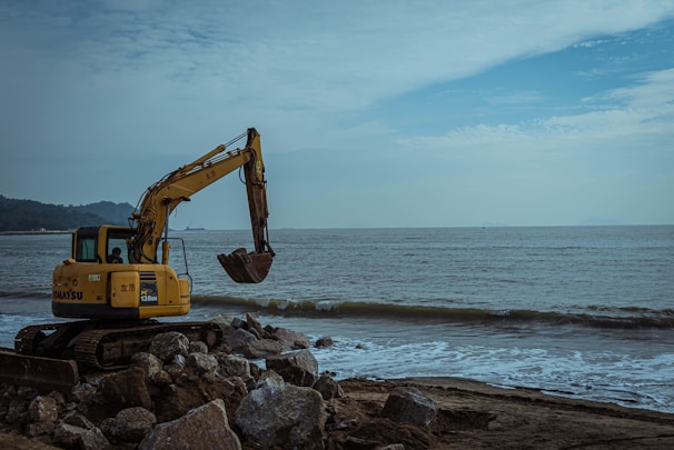 A yellow excavator is positioned on a rocky shoreline next to the ocean. The machine sits atop large stones, with its arm and bucket extended. The sea is calm, with small waves gently reaching the shore, and the sky is partly cloudy with patches of blue.