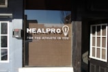 A storefront window with the logo 'MEALPRO' and the tagline 'FOR THE ATHLETE IN YOU' displayed prominently. There is a lightbulb graphic containing a fork inside it next to the logo. Surrounding the window, there are sections of a building facade including parts of two adjacent doors.