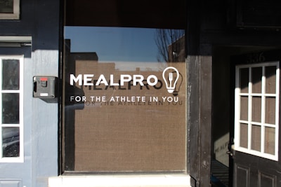 A storefront window with the logo 'MEALPRO' and the tagline 'FOR THE ATHLETE IN YOU' displayed prominently. There is a lightbulb graphic containing a fork inside it next to the logo. Surrounding the window, there are sections of a building facade including parts of two adjacent doors.