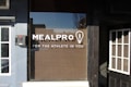 A storefront window with the logo 'MEALPRO' and the tagline 'FOR THE ATHLETE IN YOU' displayed prominently. There is a lightbulb graphic containing a fork inside it next to the logo. Surrounding the window, there are sections of a building facade including parts of two adjacent doors.