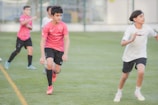 Several young boys are actively playing soccer on a grassy field. They are wearing sports attire with a mix of pink and white shirts and black shorts, and are in the midst of a soccer drill or game. Their expressions show focus and determination as they move energetically, with one boy in a pink shirt leading the action.
