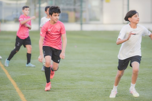 Several young boys are actively playing soccer on a grassy field. They are wearing sports attire with a mix of pink and white shirts and black shorts, and are in the midst of a soccer drill or game. Their expressions show focus and determination as they move energetically, with one boy in a pink shirt leading the action.