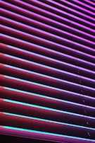 Horizontal blinds are illuminated by vibrant neon colors, creating a striking contrast between the shades of purples, pinks, and blues. The light casts colorful reflections and shadows across the blinds, giving them an abstract, modern appearance.