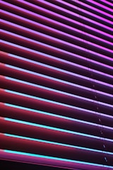 Horizontal blinds are illuminated by vibrant neon colors, creating a striking contrast between the shades of purples, pinks, and blues. The light casts colorful reflections and shadows across the blinds, giving them an abstract, modern appearance.