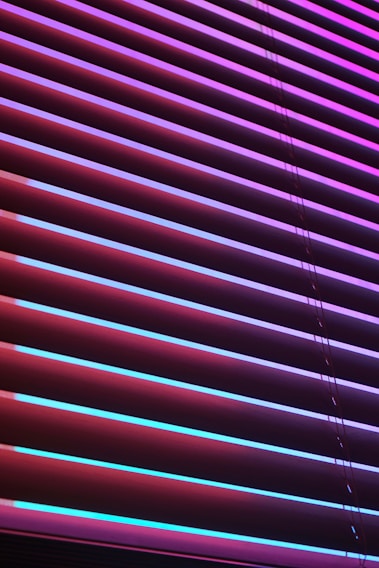 Horizontal blinds are illuminated by vibrant neon colors, creating a striking contrast between the shades of purples, pinks, and blues. The light casts colorful reflections and shadows across the blinds, giving them an abstract, modern appearance.
