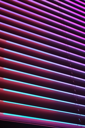 Horizontal blinds are illuminated by vibrant neon colors, creating a striking contrast between the shades of purples, pinks, and blues. The light casts colorful reflections and shadows across the blinds, giving them an abstract, modern appearance.