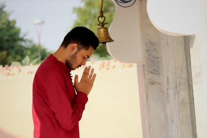 A person in a red shirt stands in a prayerful pose with hands joined in front of a bell. The individual is positioned outdoors near a white and marble structure. The background is blurred, featuring greenery and a cream-colored wall.