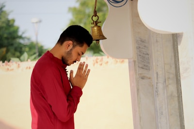 A person in a red shirt stands in a prayerful pose with hands joined in front of a bell. The individual is positioned outdoors near a white and marble structure. The background is blurred, featuring greenery and a cream-colored wall.
