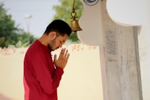 A person in a red shirt stands in a prayerful pose with hands joined in front of a bell. The individual is positioned outdoors near a white and marble structure. The background is blurred, featuring greenery and a cream-colored wall.