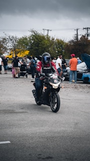 A person wearing a black helmet and a red and black jacket rides a black motorcycle in an open outdoor area. Several people are gathered in the background near parked cars, and a child in a stroller is visible. The sky is overcast, suggesting a gloomy atmosphere.