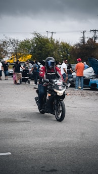 A person wearing a black helmet and a red and black jacket rides a black motorcycle in an open outdoor area. Several people are gathered in the background near parked cars, and a child in a stroller is visible. The sky is overcast, suggesting a gloomy atmosphere.