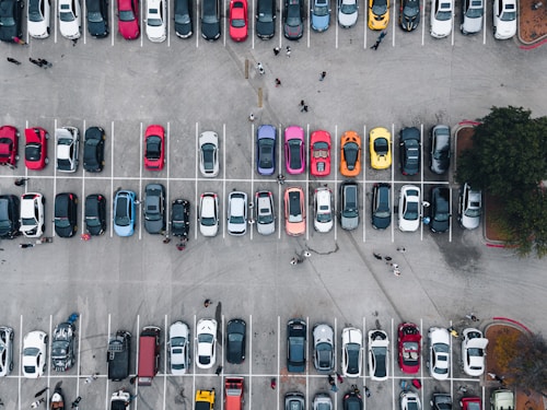An aerial view of a large parking lot filled with numerous colorful cars. The cars are neatly parked in distinct rows, with people walking along the aisles and pathways. The variety of car colors includes red, blue, white, yellow, black, and orange.