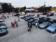 A group of people gathered in a parking lot filled with high-end sports cars, including various luxury brands in dark shades. The scene appears to be a car meet or show, with attendees chatting and inspecting the vehicles. Trees and buildings are visible in the background, with a largely overcast sky.