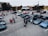 A group of people gathered in a parking lot filled with high-end sports cars, including various luxury brands in dark shades. The scene appears to be a car meet or show, with attendees chatting and inspecting the vehicles. Trees and buildings are visible in the background, with a largely overcast sky.