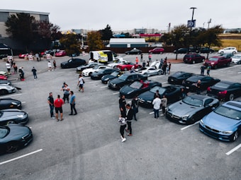 A group of people gathered in a parking lot filled with high-end sports cars, including various luxury brands in dark shades. The scene appears to be a car meet or show, with attendees chatting and inspecting the vehicles. Trees and buildings are visible in the background, with a largely overcast sky.