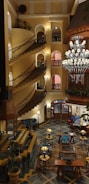 A grand hotel interior featuring a spiral staircase with ornate railings to the left side and a large, decorative chandelier hanging from the ceiling. The walls are adorned with arched windows and ambient lighting. The floor exhibits a geometric pattern with comfortable seating arrangements scattered throughout. Potted plants are placed strategically, adding a touch of greenery.