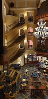 A grand hotel interior featuring a spiral staircase with ornate railings to the left side and a large, decorative chandelier hanging from the ceiling. The walls are adorned with arched windows and ambient lighting. The floor exhibits a geometric pattern with comfortable seating arrangements scattered throughout. Potted plants are placed strategically, adding a touch of greenery.