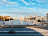 An urban river scene with modern buildings on the right and older, more residential structures with trees on the left. Construction cranes are visible, indicating ongoing development. The sky is partly cloudy with a few patches of blue, and the water reflects the surrounding architecture.