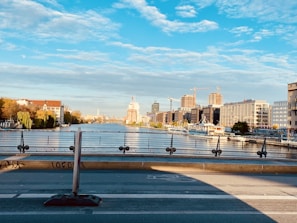 An urban river scene with modern buildings on the right and older, more residential structures with trees on the left. Construction cranes are visible, indicating ongoing development. The sky is partly cloudy with a few patches of blue, and the water reflects the surrounding architecture.