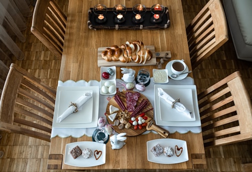 A wooden dining table is set for a meal, featuring neatly arranged place settings with white plates and napkins secured with decorative holders. A variety of foods, including sliced bread, cold cuts, cheeses, cherry tomatoes, and boiled eggs, are presented centrally on a round wooden board. Decorative elements such as painted eggs and small white bunnies add a festive touch. A row of lit candles at one end of the table creates a cozy ambiance.