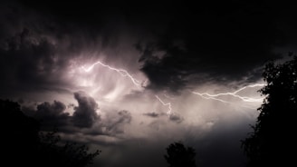 Dark stormy sky with dramatic, swirling clouds and bright streaks of lightning cutting across the scene. Silhouettes of trees are visible in the foreground, contrasting against the illuminated sky.