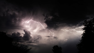 Dark stormy sky with dramatic, swirling clouds and bright streaks of lightning cutting across the scene. Silhouettes of trees are visible in the foreground, contrasting against the illuminated sky.