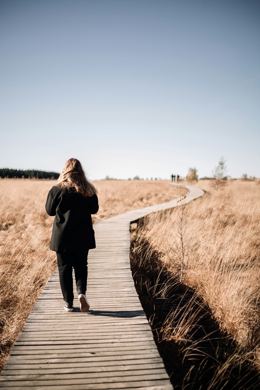 woman in black coat walking on brown wooden pathway during daytime