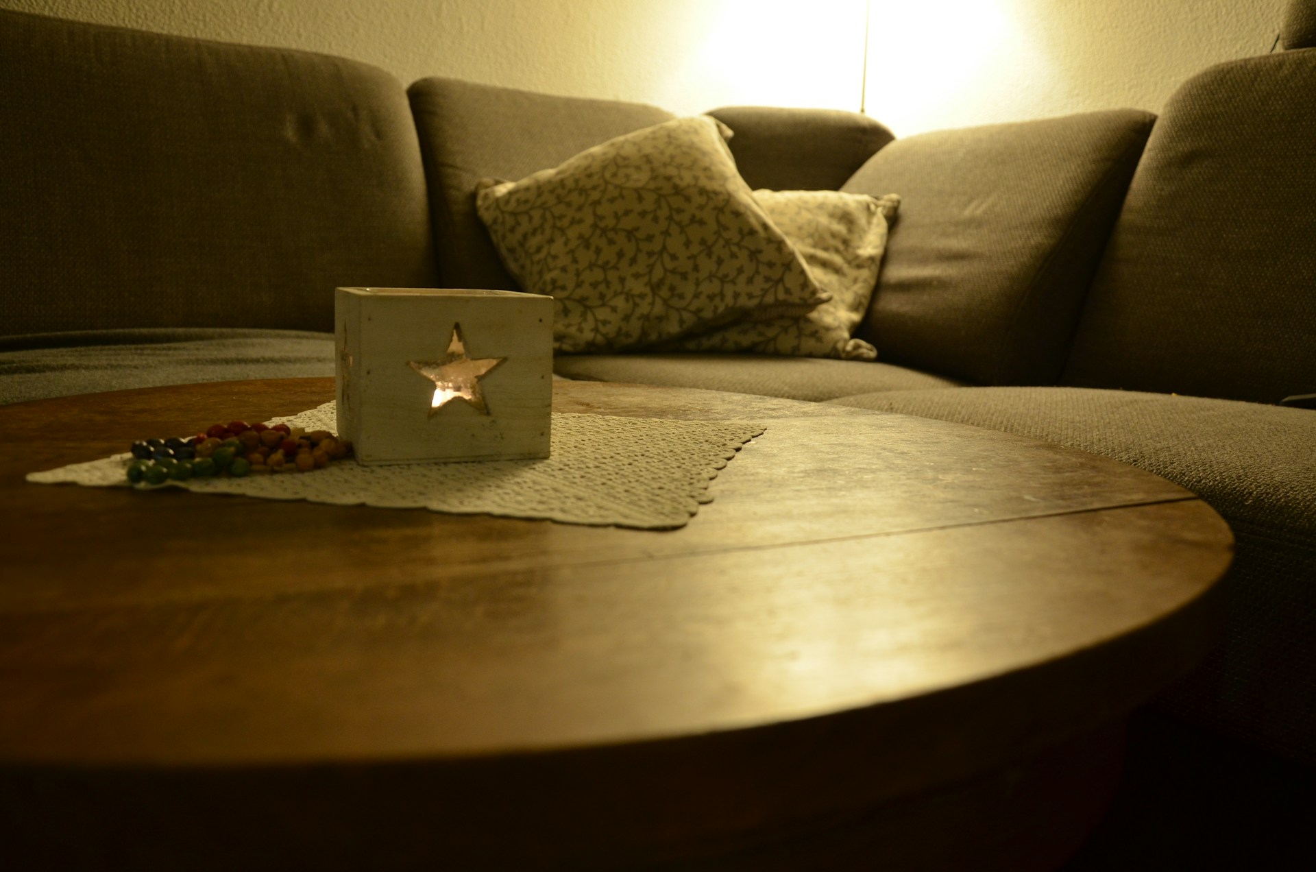 A cozy living room setting featuring a wooden round coffee table with a white lace mat on top. There is a lit wooden lantern with a star-shaped cutout and a string of colorful beads beside it. In the background, a gray upholstered sectional sofa is adorned with patterned throw pillows, and a warm lamp light softly illuminates the space.