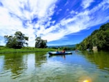 A group of people kayaking on a calm river surrounded by lush greenery and hills. The sky is clear with scattered white clouds, adding to the serene and picturesque landscape.