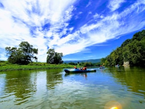 A group of people kayaking on a calm river surrounded by lush greenery and hills. The sky is clear with scattered white clouds, adding to the serene and picturesque landscape.